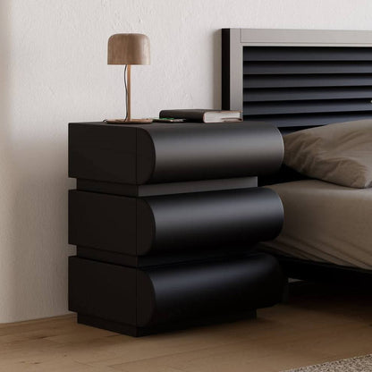 Black nightstand with a lamp and books next to a bed in a bedroom.