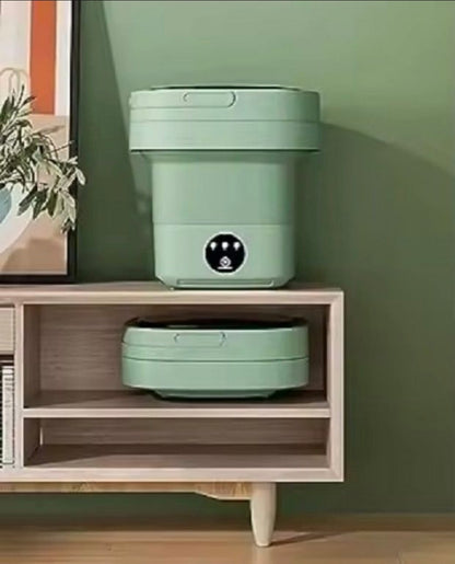 A light sage green robotic vacuum cleaner shown in two states: one upright on top of a wooden shelf and one lying flat underneath, set against a sage green wall.