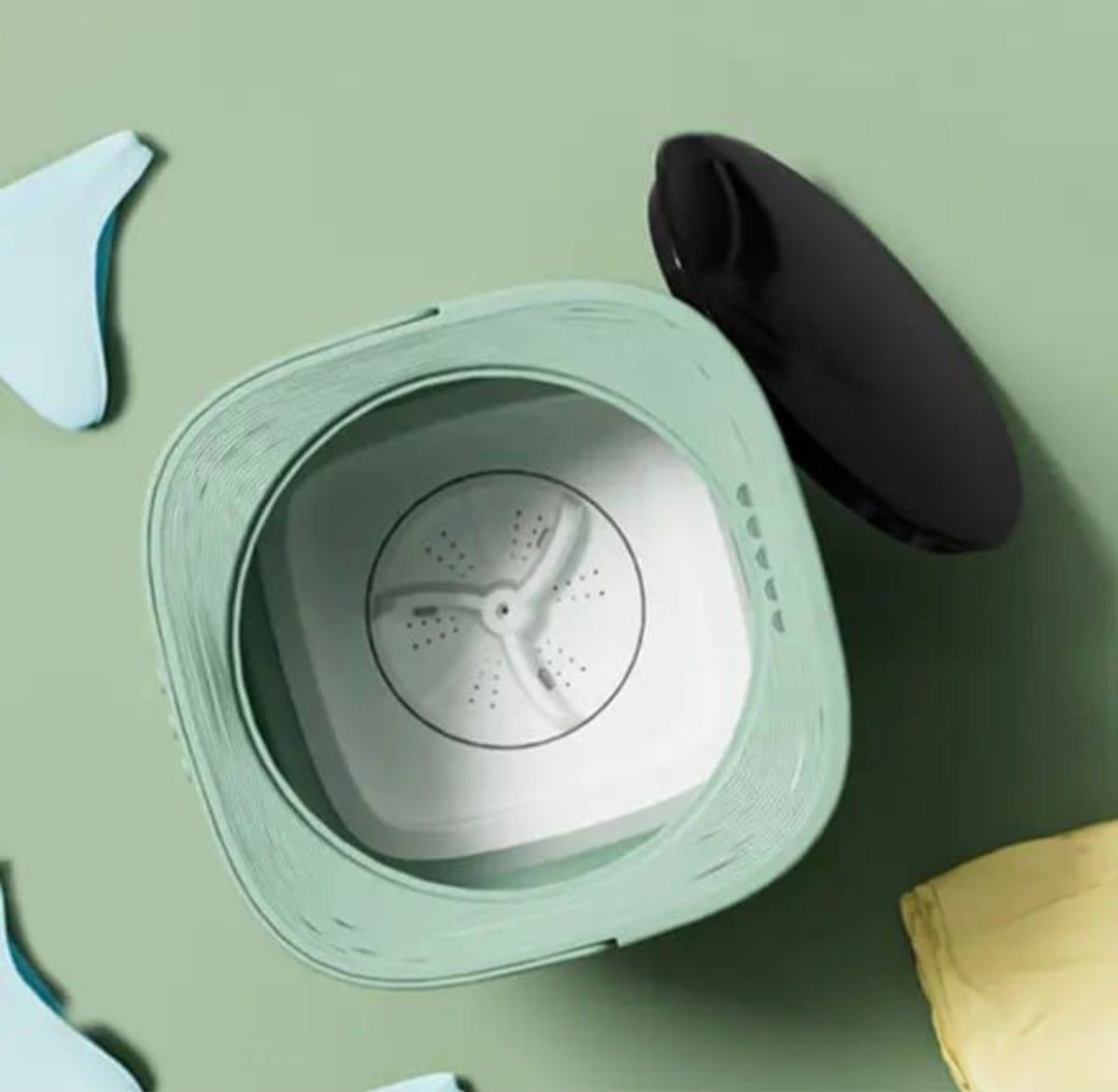 A top-down view of a compact mini washing machine with a pale sage green body, white interior drum, and a detached black lid placed beside it on a matching sage green background.