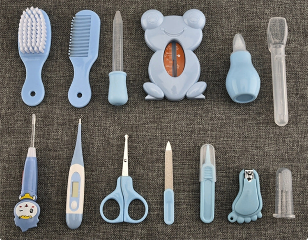 A set of baby grooming tools arranged on a textured gray fabric background, including a brush, comb, nail clipper, thermometer, scissors, nail file, and other light blue and clear plastic grooming accessories.